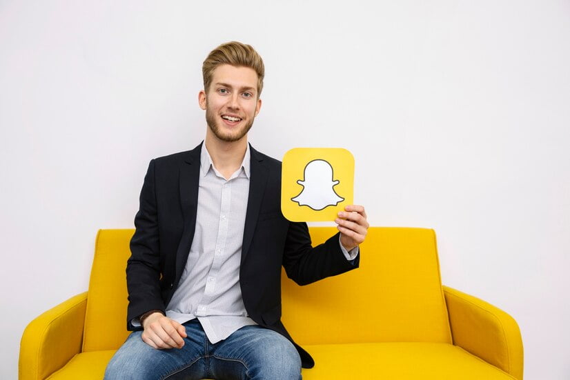 تسجيل الدخول لسناب شات برقم الجوال - انشاء حساب Snapchat 3 portrait young man sitting yellow sofa holding snapchat icon 23 2147847510